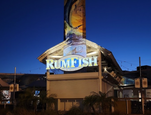 Sign for RumFish Grill illuminated at dusk, featuring a colorful mural and palm trees in the foreground.