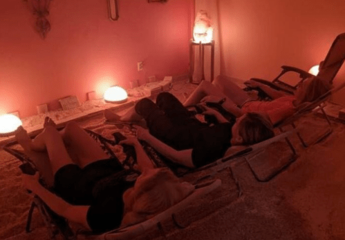 Three people relax on lounge chairs in a softly lit room with pink walls and glowing salt lamps.