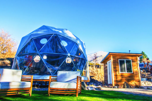 A geodesic dome with large windows, surrounded by green grass and wooden seating, next to a small wooden cabin.