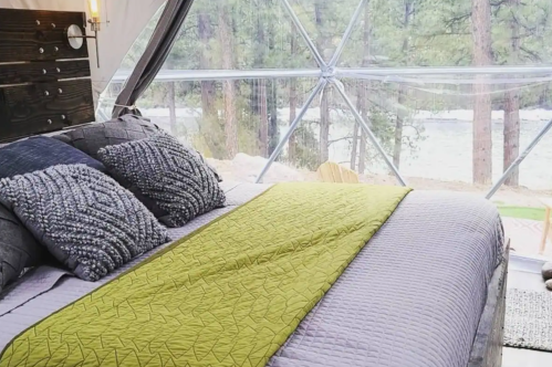 Cozy bedroom in a geodesic dome with a green blanket, textured pillows, and a view of trees outside.