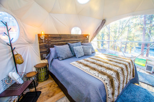Cozy interior of a geodesic dome bedroom with a bed, decorative blankets, and large windows overlooking nature.