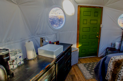 Interior of a cozy yurt with a green door, sink, and wooden flooring, featuring circular windows and simple decor.