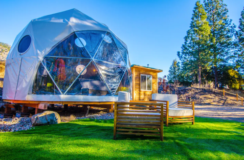 A geodesic dome structure with large windows, surrounded by greenery and a wooden seating area in a scenic outdoor setting.