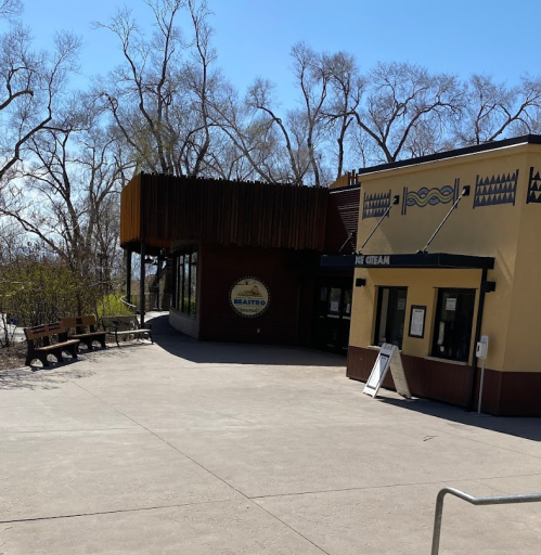 A modern building with large windows and a wooden overhang, surrounded by trees and a paved walkway.