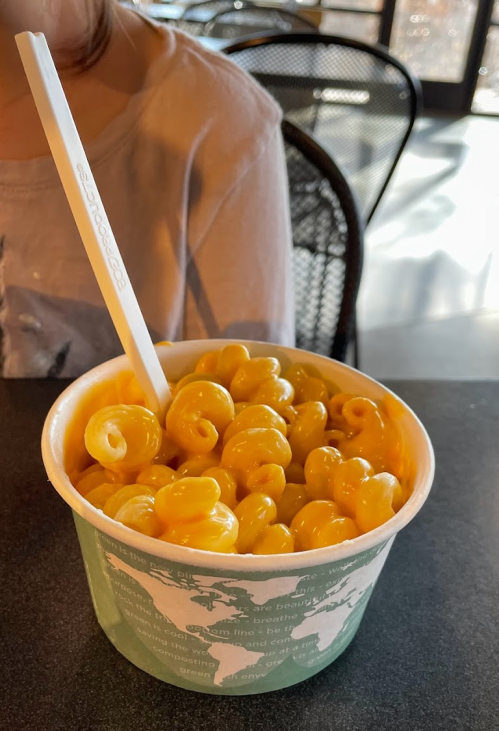 A cup of macaroni and cheese with spiral pasta, sitting on a table in a casual dining setting.