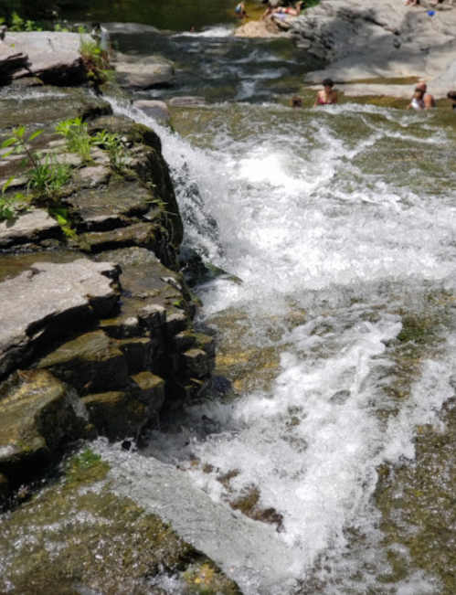 A cascading waterfall flows over rocky terrain, with people enjoying the water in the background.