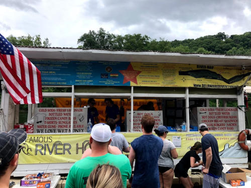 A busy food stand at a river with people ordering, featuring a menu of hot dogs, drinks, and snacks. An American flag is visible.