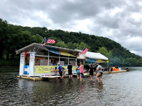 A group of people wade in water near a floating restaurant, surrounded by greenery and cloudy skies. American flags are visible.
