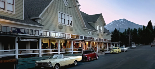 Historic buildings line a street with classic cars parked outside, set against a backdrop of mountains at dusk.
