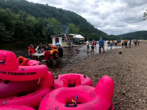 A river scene with people enjoying tubing, a food stand, and colorful inner tubes on the shore under a cloudy sky.
