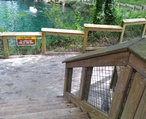 A wooden staircase leads down to a pond with a caution sign warning about swimming at one's own risk.