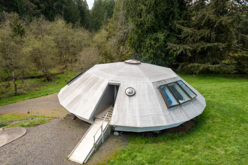 A unique, round concrete house with a sloped roof, surrounded by trees and grass, featuring large windows and a walkway.
