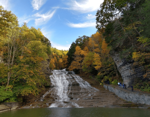 A scenic waterfall surrounded by autumn foliage and a clear blue sky, with people walking along the shore.