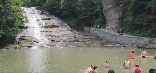 A scenic waterfall cascades into a swimming area, with people enjoying the water and walking along a nearby path.