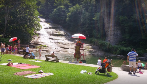 A sunny park scene with people enjoying a waterfall, lounging on blankets, and a lifeguard station by the water.