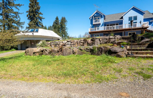 A blue house with a deck sits beside a rocky landscape and a unique, modern structure under a clear blue sky.