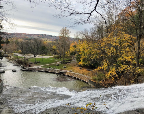 A serene view of a waterfall cascading into a river, surrounded by autumn trees and a park with benches.