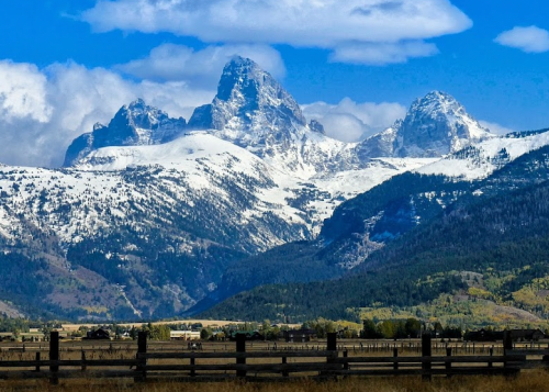 Snow-capped mountains rise majestically under a blue sky, with green valleys and a wooden fence in the foreground.