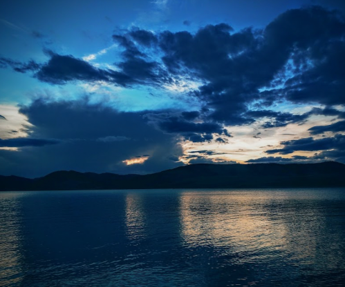 A serene lake at dusk, reflecting dark clouds and a blue sky, with mountains in the background.