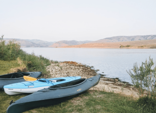 Two kayaks on the shore of a calm lake, surrounded by greenery and distant hills under a clear sky.