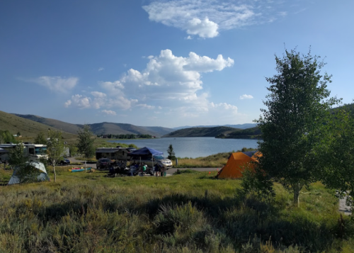 A serene lakeside campsite with tents and RVs, surrounded by hills and scattered clouds in a blue sky.