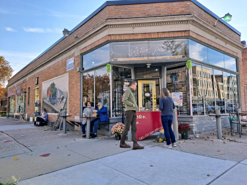 A cozy bakery storefront with people outside, enjoying the day and chatting, surrounded by autumn decorations.