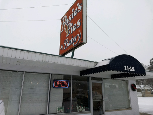 A snowy scene of Rosie's Pies & Bakery, featuring an "OPEN" sign and a large outdoor sign.
