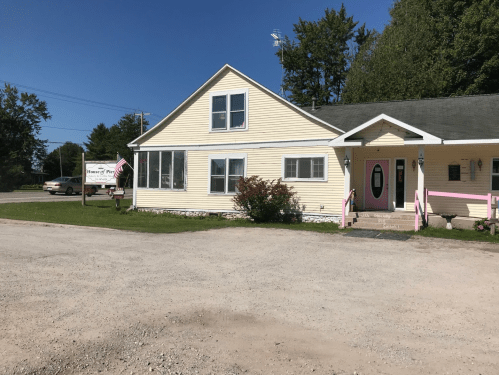 A yellow house with a pink door and sign, surrounded by trees and a gravel driveway under a clear blue sky.
