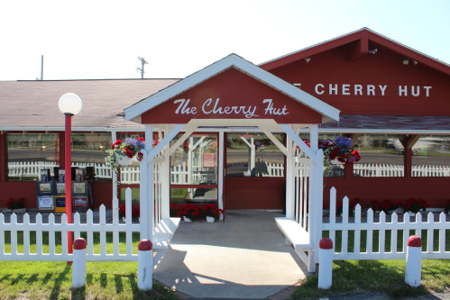 A red building with a white picket fence, featuring a sign that reads "The Cherry Hut" and colorful flower pots.