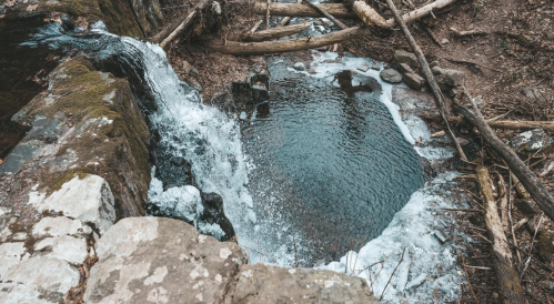 Aerial view of a rocky waterfall flowing into a deep, icy blue pool surrounded by fallen branches.