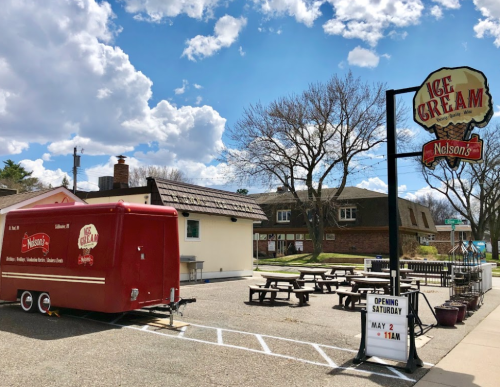 A red ice cream truck parked outside with picnic tables, under a blue sky with clouds, announcing an opening date.