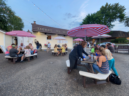 A busy ice cream shop with striped umbrellas, people enjoying treats at picnic tables during twilight.