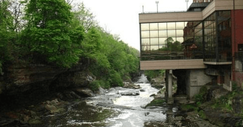 A modern building with large windows overlooks a flowing river surrounded by lush greenery and rocky banks.