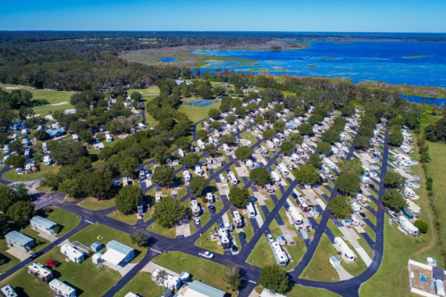 Aerial view of a large RV park with numerous parked RVs, surrounded by greenery and a blue lake in the background.