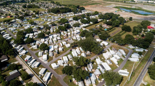 Aerial view of a mobile home park with numerous trailers and green spaces, surrounded by trees and open land.