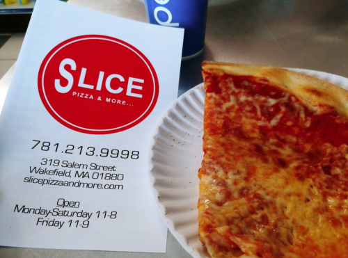 A slice of cheese pizza on a paper plate next to a menu for Slice Pizza & More in Wakefield, MA.
