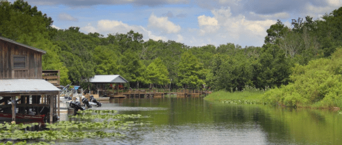 A serene waterway lined with greenery, featuring boats docked at wooden structures under a blue sky with clouds.