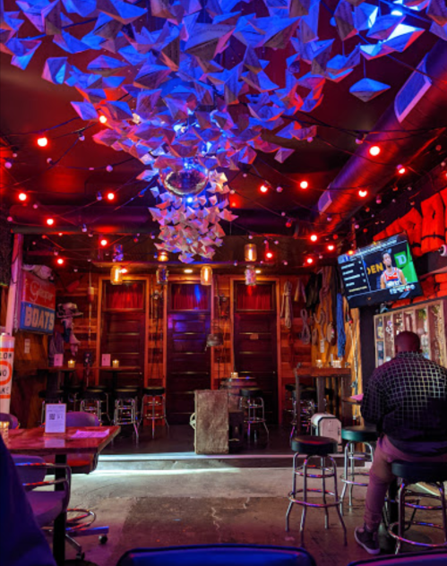 A vibrant bar interior with colorful lights, paper decorations, and a disco ball, featuring empty stools and a TV screen.