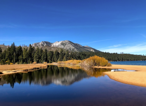 A serene lake surrounded by trees and mountains, with clear blue skies and autumn foliage reflected in the water.