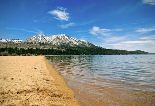 A sandy beach meets a calm lake, with snow-capped mountains and a clear blue sky in the background.