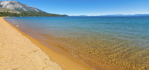 A serene beach with golden sand and clear blue water, surrounded by mountains under a clear sky.