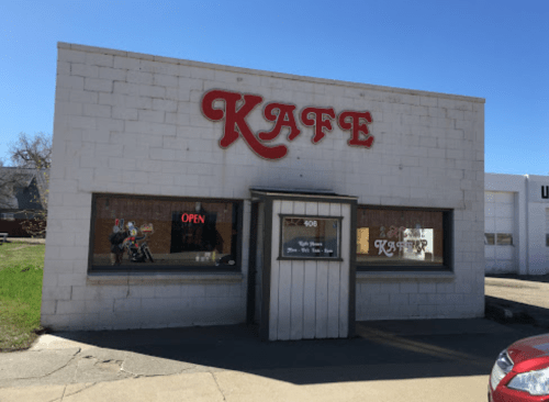 A small, white building with a red "KAFE" sign and an "OPEN" sign in the window, set against a clear blue sky.