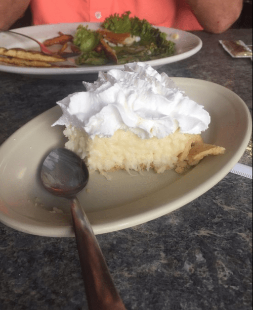 A slice of coconut cream pie topped with whipped cream on a plate, with a spoon beside it.