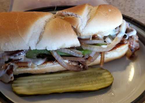 A sandwich filled with grilled onions, green peppers, and meat, served with a pickle on the side.