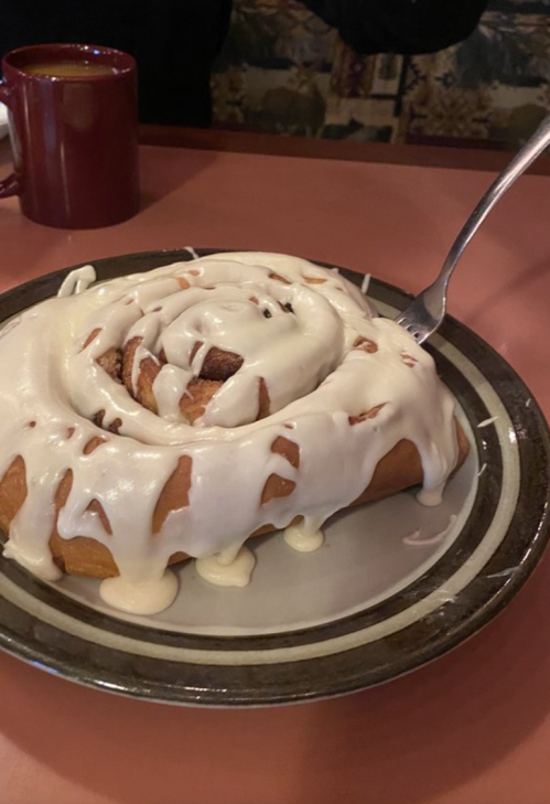 A large cinnamon roll topped with creamy icing on a plate, with a fork beside it and a mug in the background.