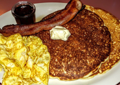 A plate of pancakes with a side of bacon, an omelet, and a small cup of syrup. Butter is melted on the pancakes.