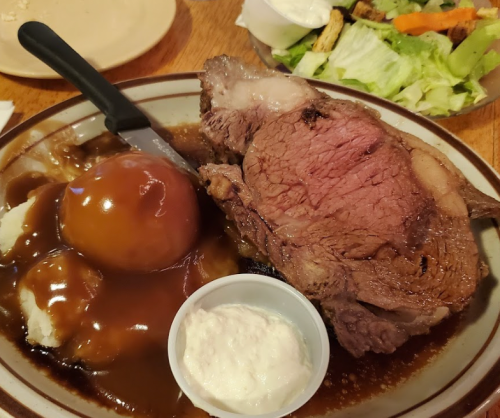 A plate featuring a large slice of prime rib with gravy, mashed potatoes, a side of applesauce, and a salad.