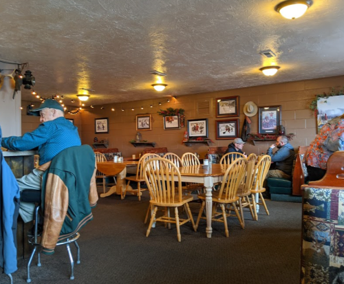 A cozy restaurant interior with wooden tables, chairs, and patrons seated at tables, decorated with framed pictures.