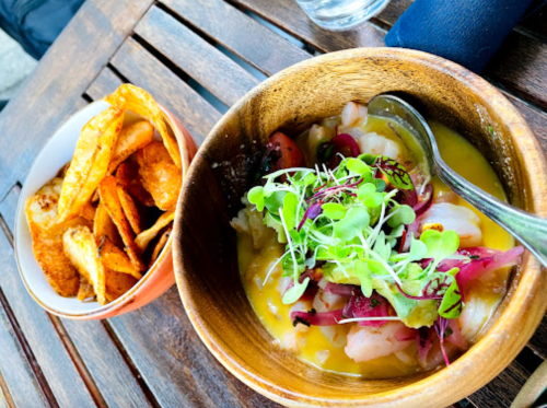A wooden bowl with colorful ceviche topped with microgreens, alongside a small bowl of crispy chips on a wooden table.
