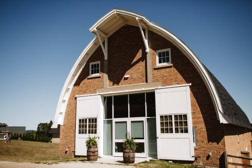 A large, red brick barn with a curved roof, featuring white doors and windows, set against a clear blue sky.